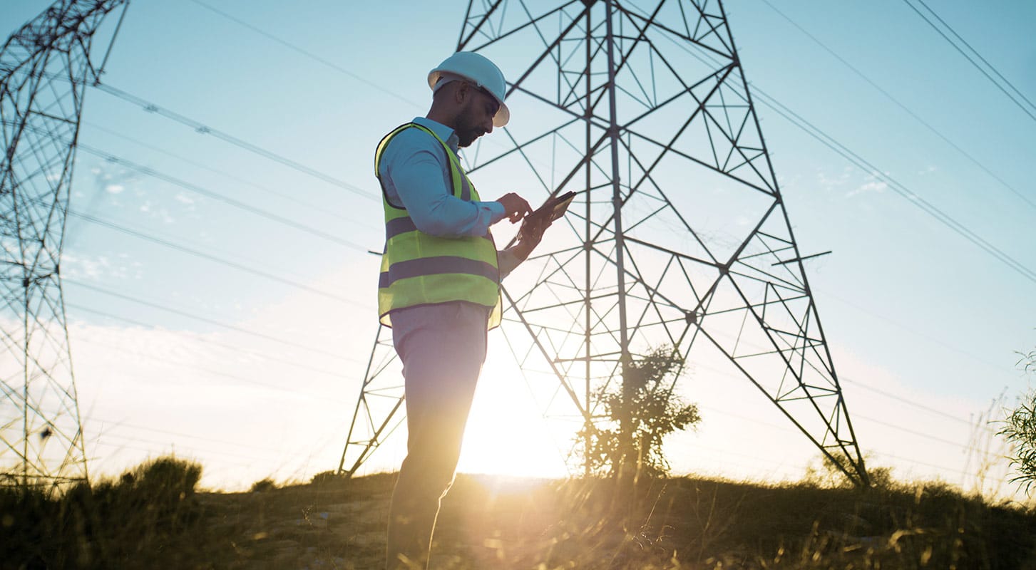 A man in a hard hat stands beside a tall electricity tower, surveying the area for maintenance work.